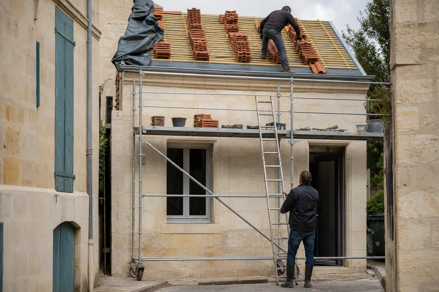 Échafaudage dressé contre une façade en pierre calcaire, tuiles empilées sur les liteaux du toit